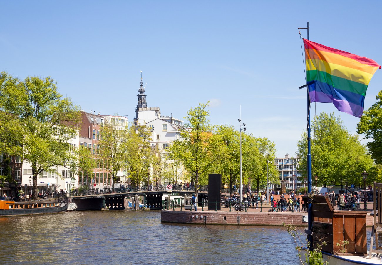 A rainbow pride flag flies by a canal in a city with trees, buildings, and a bridge under a clear blue sky.