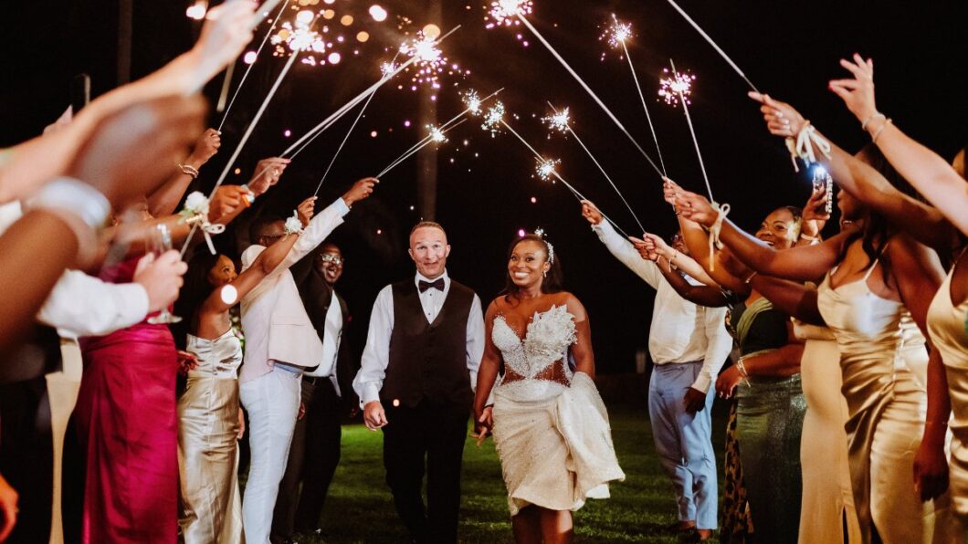 affordable guest travel - featured image A bride and groom walk hand in hand under a tunnel of sparklers held by guests at an outdoor nighttime celebration.
