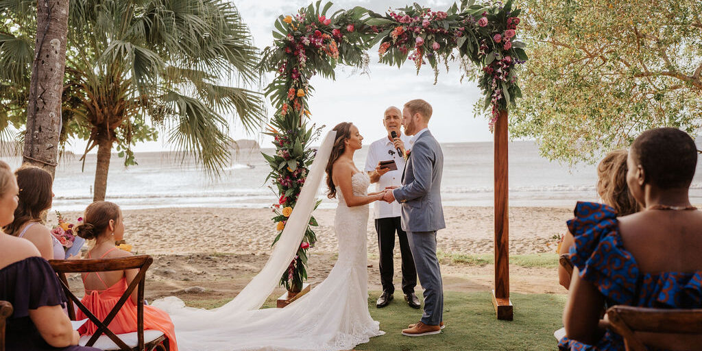 Alexandria_Janssen_Estefanía Herrera Photography_A+JC-160 A bride and groom exchange vows under a floral arch at a beachside wedding ceremony, with guests seated nearby and the ocean in the background.