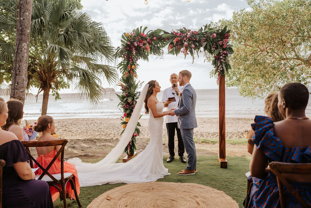 A bride and groom exchange vows under a floral arch at a beachside wedding ceremony, with guests seated nearby and the ocean in the background.