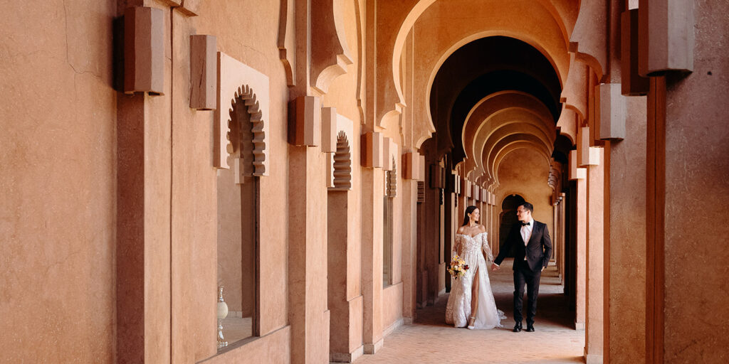 Marrakech - AMV retreat A couple dressed in formal wedding attire walks hand in hand through a corridor with arches and warm-toned walls.