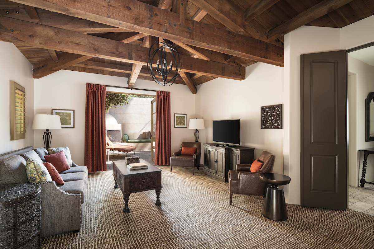 Living room with exposed wooden beams, neutral furniture, large windows with red curtains, a TV on a cabinet, and a view of an outdoor fireplace.