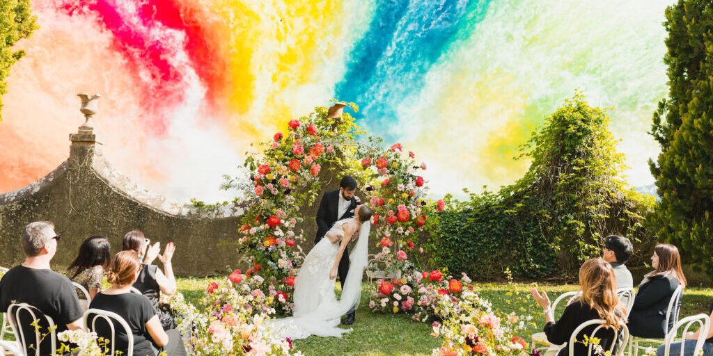 Borgo Castelvecchi Editorial Photo Shoot A bride and groom kiss under a floral arch at an outdoor wedding ceremony, with guests seated among flowers and a vibrant, colorful smoke fireworks in the background.