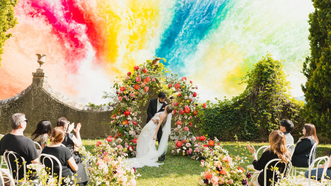 Borgo Castelvecchi Editorial Photo Shoot A bride and groom kiss under a floral arch at an outdoor wedding ceremony, with guests seated among flowers and a vibrant, colorful smoke fireworks in the background.