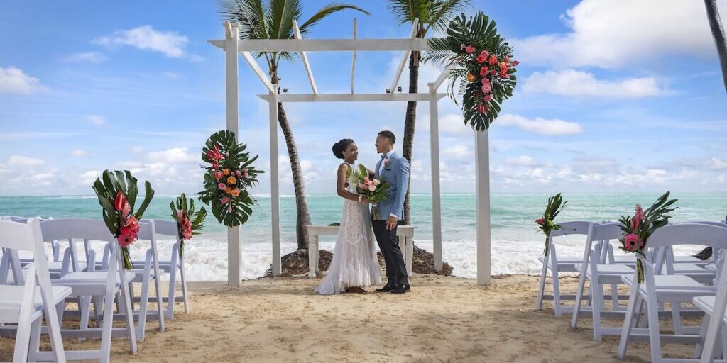 grand-palladium-punta-cana1 Bride and groom stand under a decorated white pergola on a sandy beach with palm trees, ocean, and flower arrangements in the background.