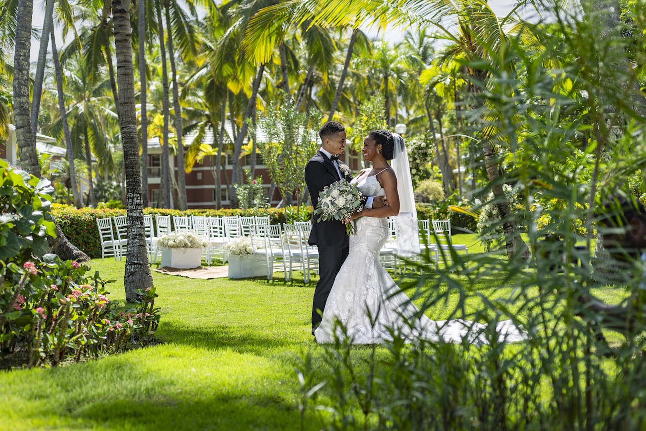 A bride and groom stand together outdoors in a tropical garden, surrounded by greenery and palm trees, with white chairs set up in the background.