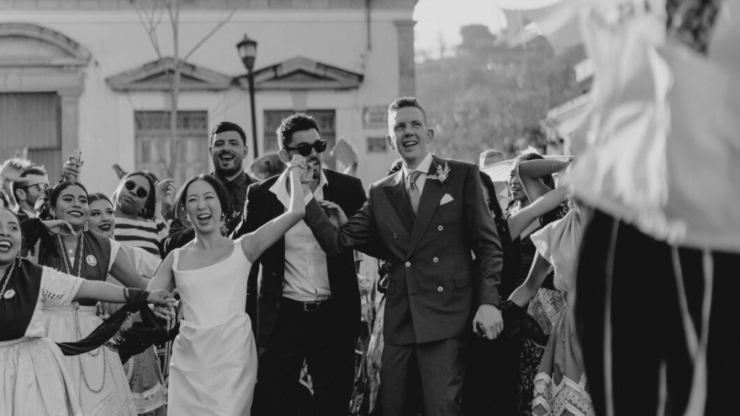Guest Communications - Featured Image A black and white image of a wedding procession with a joyful bride and groom holding hands, surrounded by smiling, formally dressed guests and performers in traditional attire.
