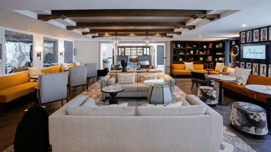 hotel-amarano-featured-image Spacious living room with beige sofas, armchairs, a glass coffee table, wooden ceiling beams, bookshelves, and a yellow couch.