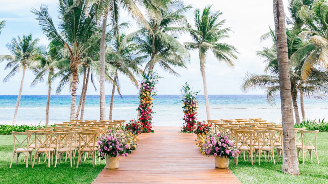 info@bohemiadelmar.com A wooden aisle lined with floral arrangements and chairs leads to two colorful flower pillars on a beach, with palm trees and the ocean in the background.