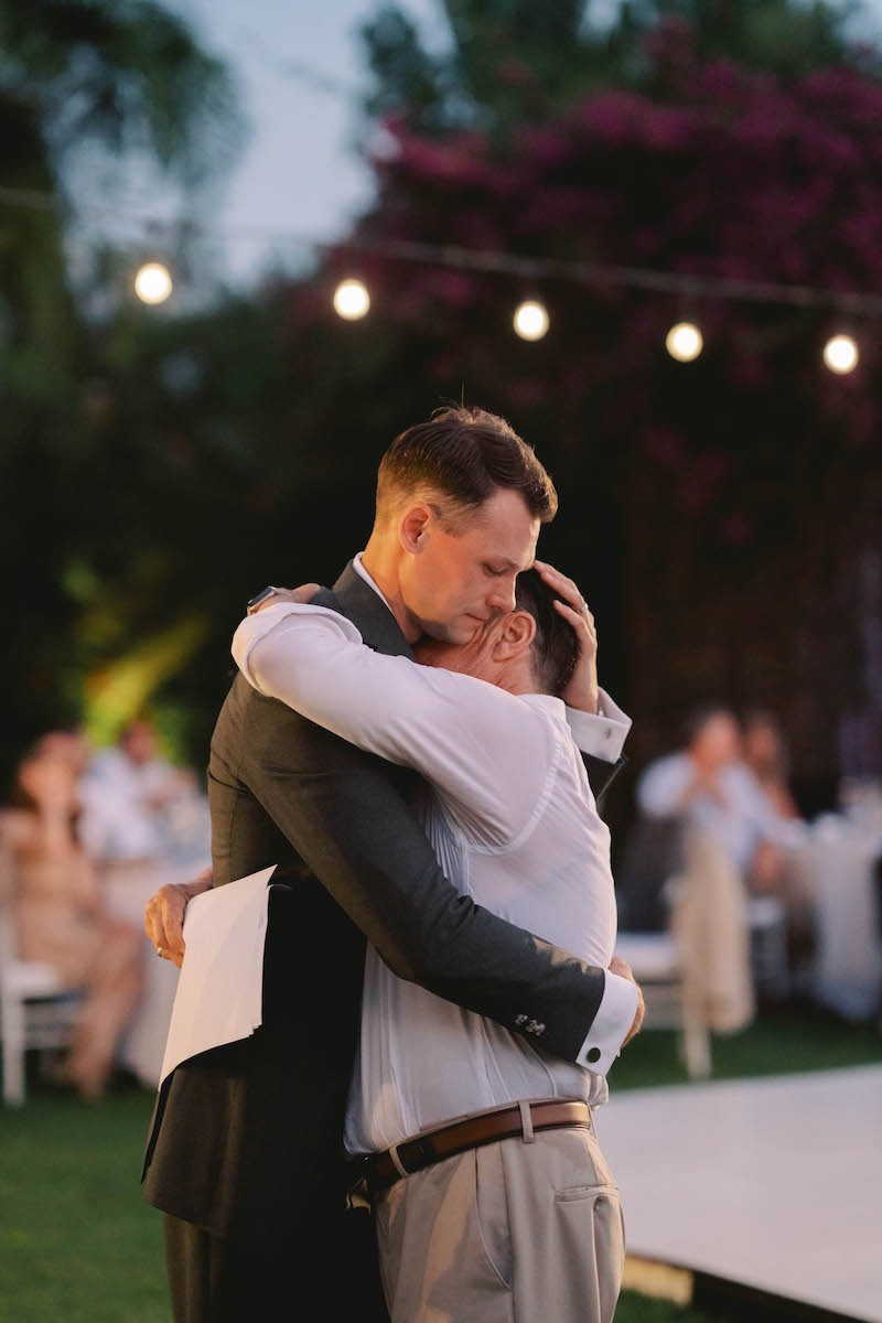 Two men embrace at an outdoor event, one holding a piece of paper. String lights and seated guests are visible in the background.