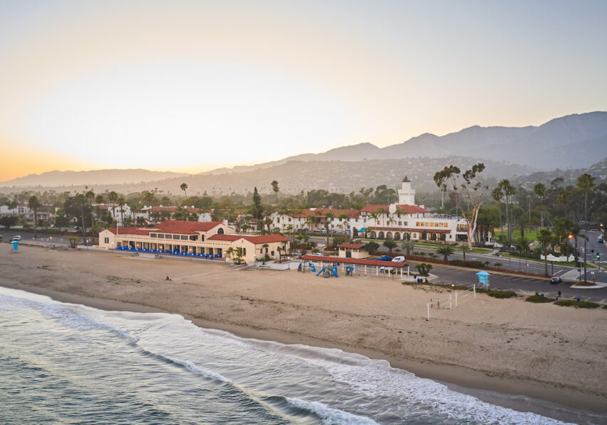 Aerial view of a beach at sunset with a coastal town, palm trees, and mountains in the background.