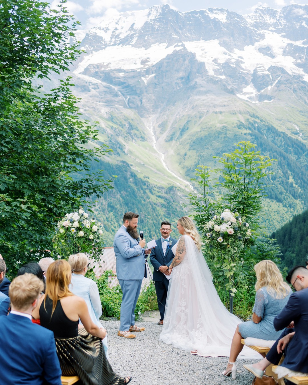 A couple exchanges vows outdoors on a mountain overlook, surrounded by guests, greenery, and floral arrangements, with snow-capped peaks in the background.