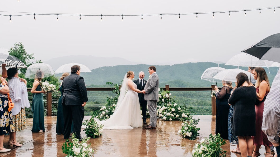 Morgan & Austin's Wedding A bride and groom stand at an outdoor wedding ceremony under string lights, holding hands, while guests observe with umbrellas on a rainy day. The background shows a foggy mountain landscape.