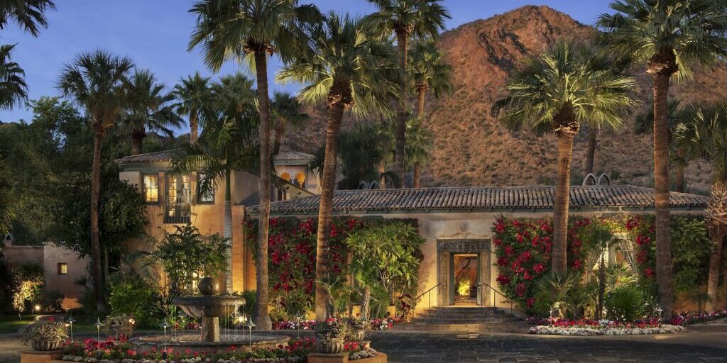 Mark Boisclair Photography, Inc. A Mediterranean-style villa with palm trees, flowering vines, and a courtyard fountain sits at the base of a rocky mountain at dusk.
