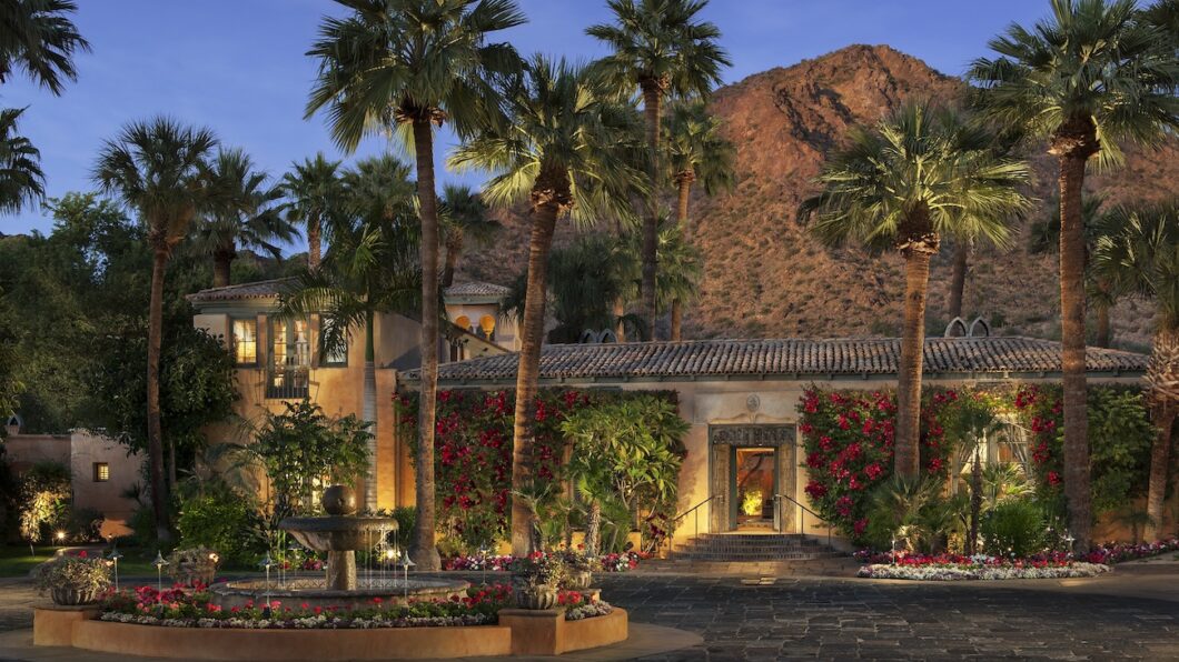Mark Boisclair Photography, Inc. A Mediterranean-style villa with palm trees, flowering vines, and a courtyard fountain sits at the base of a rocky mountain at dusk.