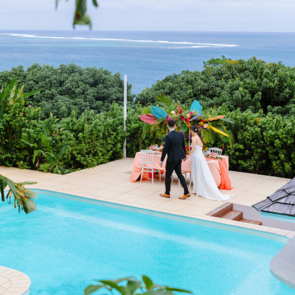 A couple stands by a decorated table near a pool overlooking the ocean in Tahiti, surrounded by lush greenery.