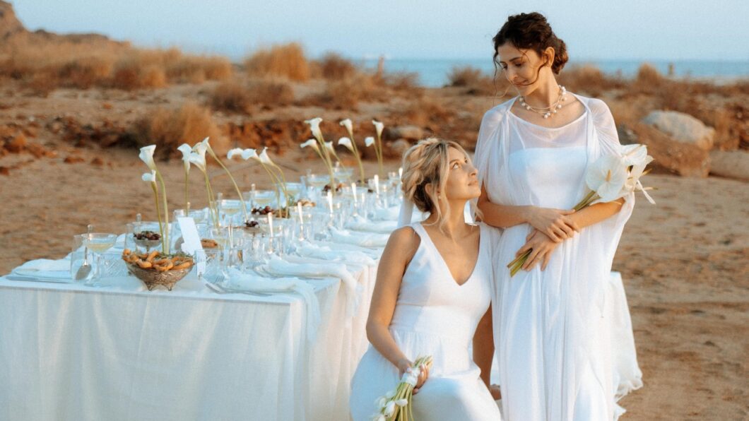 Top LGBTQ-Friendly Cities for Weddings Outside of the U.S. Two women in white dresses pose beside an outdoor table set with white flowers and tableware on a beach, with dry grass and the sea in the background.