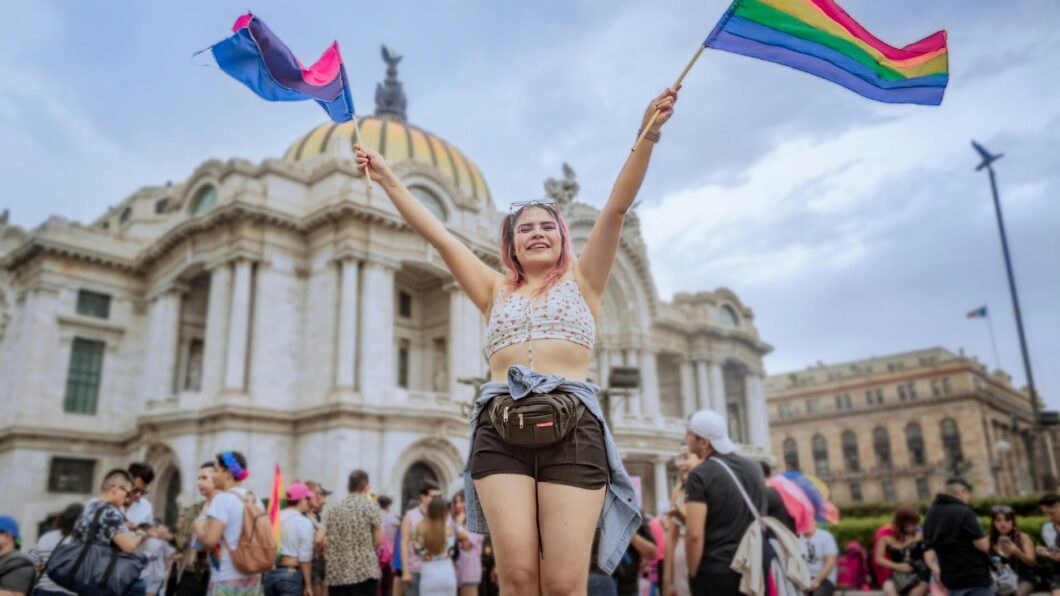 A person stands in front of a historic building, holding up two pride flags, surrounded by a crowd during a public event.