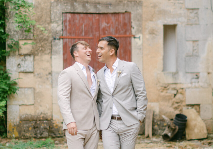 Noel and Daniel laugh together walking shoulder to shoulder away from an old French countryside building with a red door