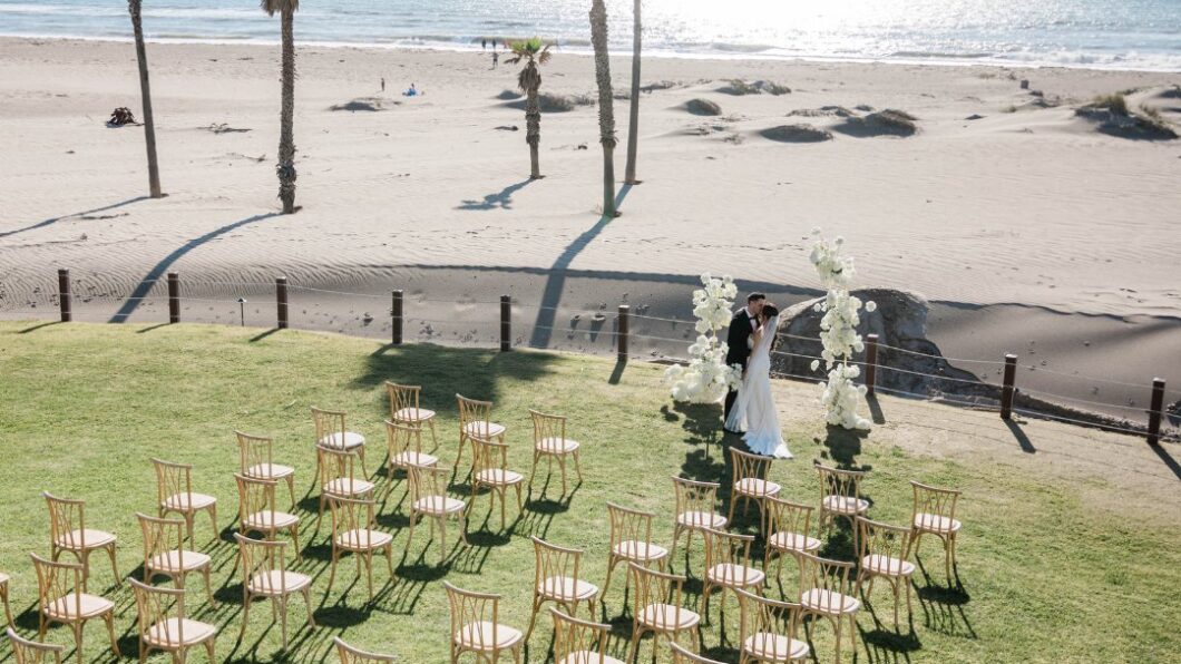 zacharidunes-visit3 A couple stands under a floral wedding arch on a grassy area near the beach, with empty chairs arranged in rows and palm trees along the shoreline.