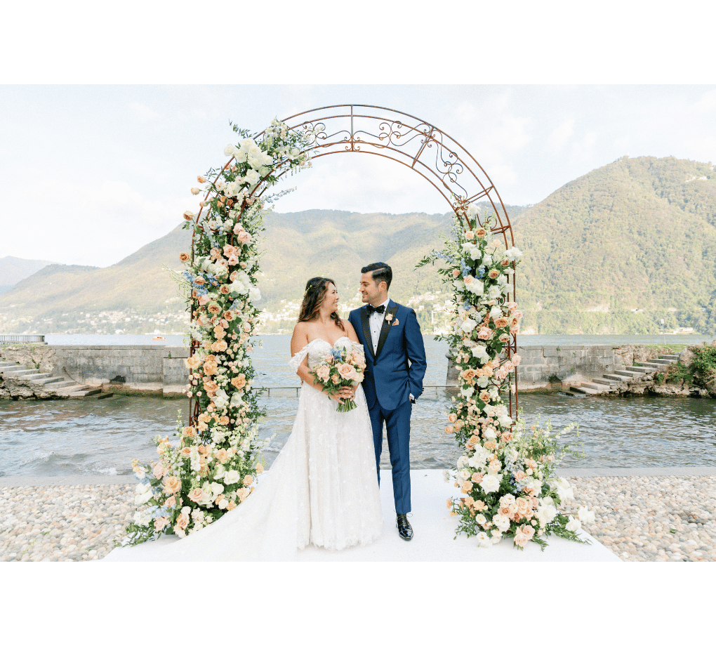 A bride and groom stand under a floral arch by a lakeside, with mountains in the background.